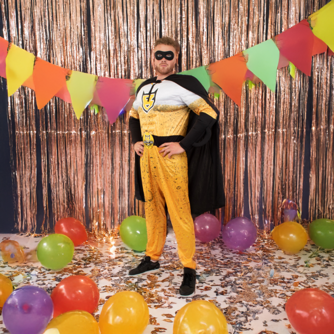 Man dressed in a beer costume, standing in a lively carnival or party setting, with colorful decorations, balloons, and confetti in the background.